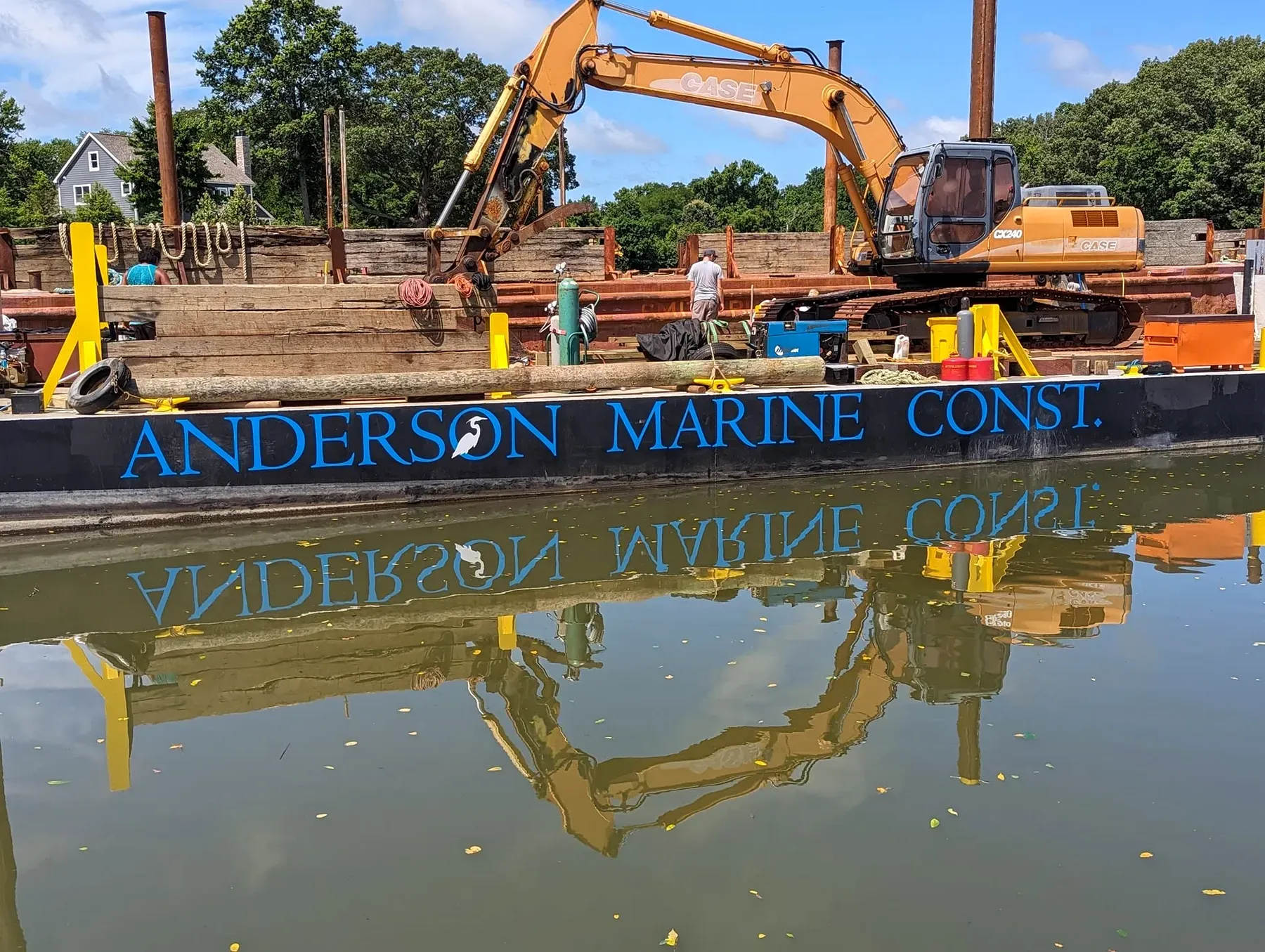 The Anderson Marine Construction barge on the job, excavator and timber pile on deck, reflected in calm water